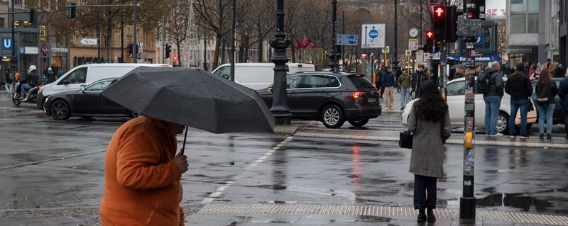 eine regnerische Straßenszene in Berlin-Charlottenburg. Eine Person mit Regenschirm geht über den Gehweg.