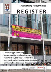 Im Hintergrund ist das Sozialamt zu sehen, an dessen Fassade ein großes Transparent in lila und gelb angebracht ist. Darauf steht Friedrichshain-Kreuzberg steht auf gegen Rassismus.