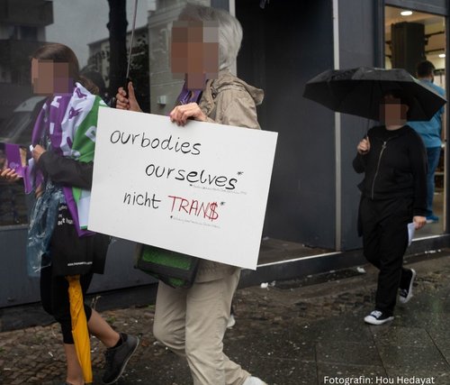 Drei Frauen. Die Gesichter sind verpixelt. Die vorderste Frau hält ein Schild mit den Worten: ourbodies ourselves - nicht tran$. Die Frau hinten rechts hält einen Regenschirm. Die Frau hinten links hat eine Fahne in lila, weiß, grün um sich.