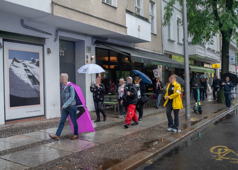 Eine Straße und ein Gehweg. Auf dem Gehweg laufen 11 Menschen im Regen. Einige tragen Regenschirme. Im Hintergrund sind Geschäfte, ein Baum und eine Laterne.