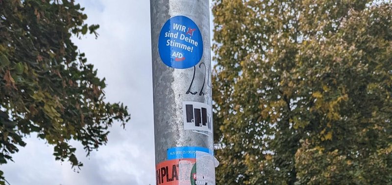 Laternenpfahl mit Bäumen im Hintergrund. Darauf sind mehrere Sticker die teilweise abgerissen sind und oben ein AfD-Aufkleber auf dem "Wir sind deine Stimme" steht.